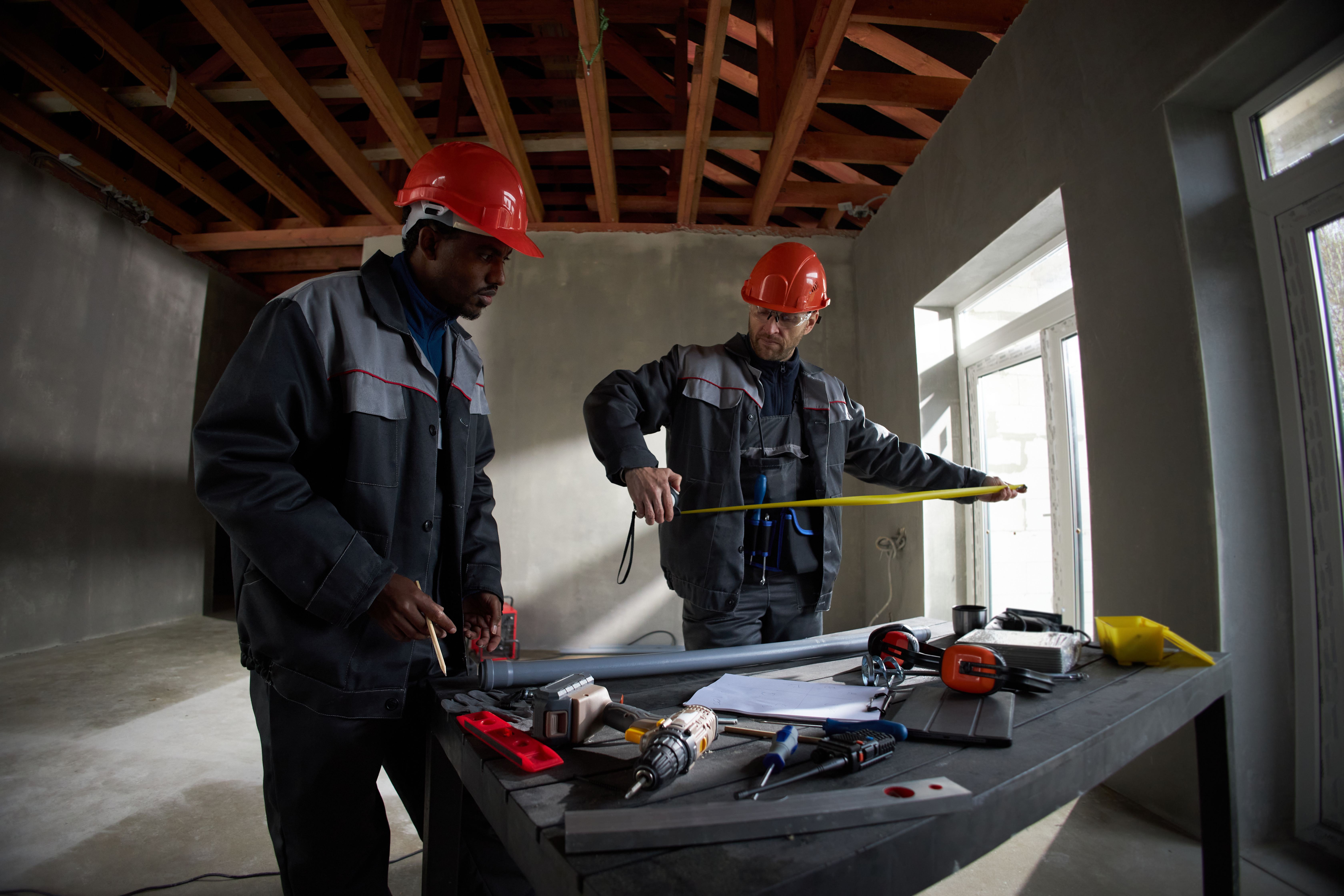 Middle aged Black man and middle aged Caucasian man wearing hard hats working together on construction site, measuring metal beam with tape measure, using tools on table